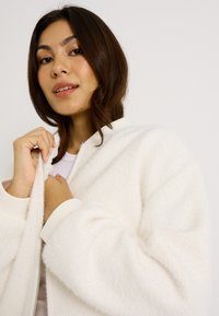 Chaqueta bomber blanca con cierre de cremallera, fabricada en un tejido texturizado, con puños acanalados y un ajuste holgado. Presenta un cuello alto y una sensación suave.