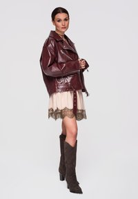 Femme portant une veste en cuir rouge foncé surdimensionnée, une robe beige clair ornée de dentelle et de hautes bottes à talons marron foncé, posant sur un fond uni.