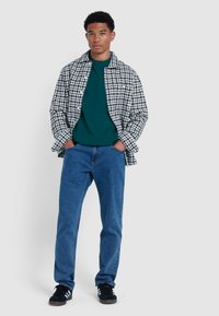 Camicia a quadri bianco e nero sopra una t-shirt verde a girocollo, jeans blu e sneakers nere con strisce bianche. Vista frontale, mani in tasca.