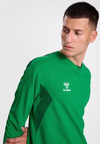 Camisetta verde a maniche lunghe realizzata in materiale morbido, con scollo a V, logo sul petto e dettagli neri a contrasto sulle spalle.