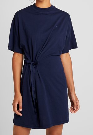 Woman wearing a navy blue short-sleeve dress with a tied knot at the waist, standing against a plain gray background.