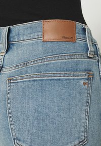 Vaqueros de mezclilla azul claro con un parche de cuero marrón etiquetado como "Madswell" en la cintura, que cuenta con cuatro bolsillos traseros y costuras de contraste.