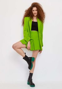 Blazer verde brillante y falda plisada a juego, combinados con botas negras hasta el tobillo con acentos verdes. Textura suave y diseño a medida.