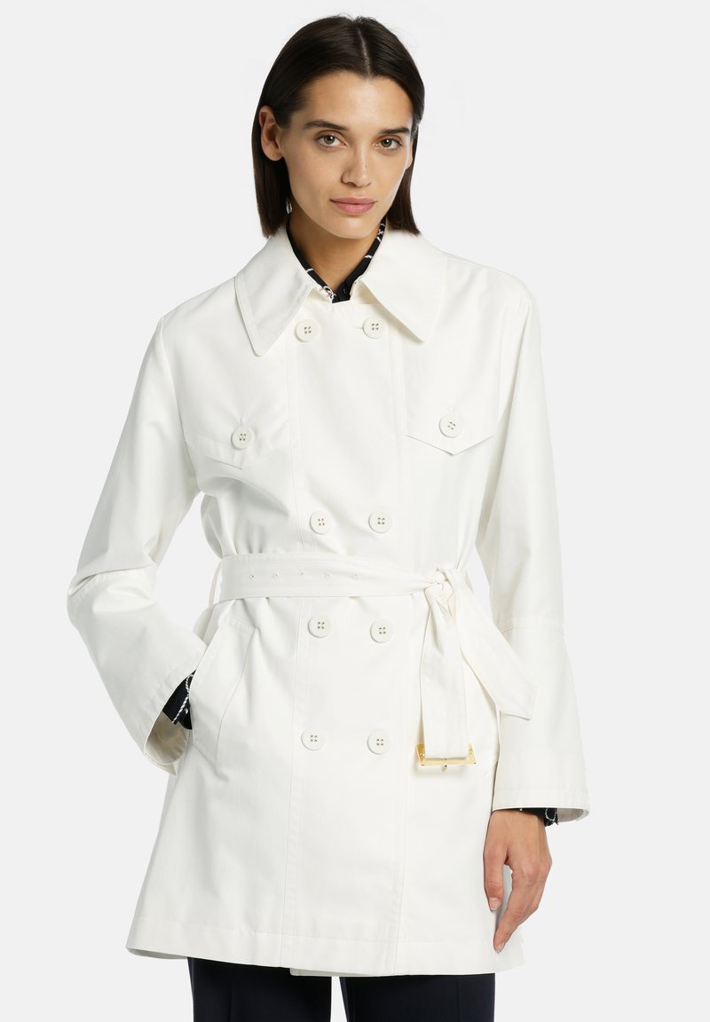 Manteau trench en coton blanc avec fermeture à double boutonnage, taille ceinturée, grand col et deux poches avant. Texture lisse.