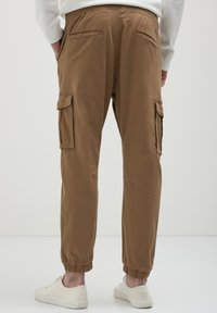 Personne portant un pantalon cargo marron avec des poignets élastiques et des poches latérales, associé à des baskets blanches et un haut à manches longues blanc.