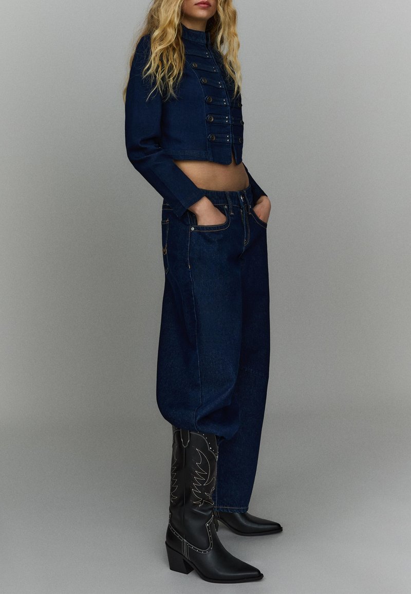 Vrouw met blond haar, gekleed in een donkerblauw kort jack, losse donkerblauwe jeans en zwarte cowboylaarzen met borduurwerk, staand met handen in de zakken.