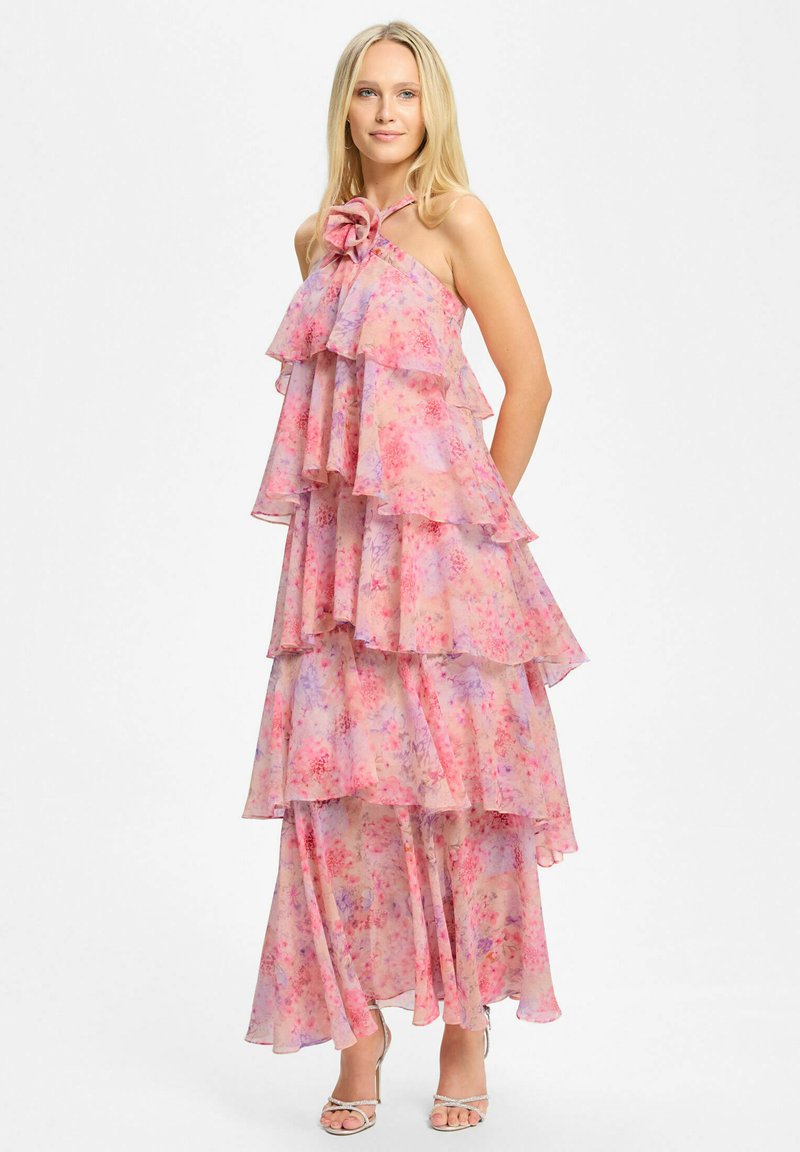 Femme blonde portant une robe bustier à volants rose à fleurs avec un grand détail de fleur au niveau du décolleté et des talons argentés à brides.