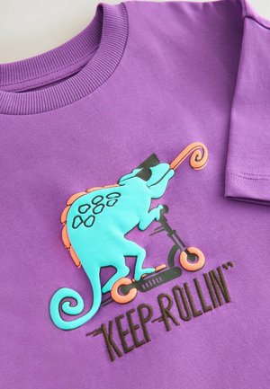 Violetti puuvillainen t-paita, jossa turkoosi kameleonttigrafiikka, joka ajaa skootterilla. Paita sisältää oransseja yksityiskohtia ja mustalla brodeeratun tekstin "KEEP ROLLIN'."