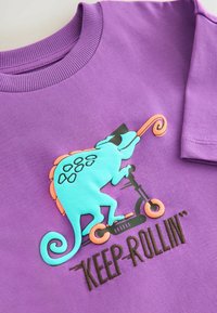 Camiseta púrpura con un gráfico de un camaleón turquesa en un scooter, con acentos naranjas. El texto dice "SIGUE RODANDO" en bordado marrón.