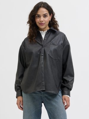 Žena s kučeravými vlasmi, oblečená v voľnej čiernej koženej košeli na gombíky cez biely top a modré džínsy, stojaca pred jednoduchým bielym pozadím.