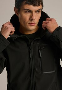 Czarna kurtka softshell z zapięciem na zamek, wysokim kołnierzem i regulowanymi mankietami. Posiada logo i kontrastowe detale zamka. Gładka tekstura.