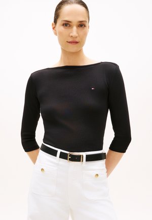 Svart, figurnära långärmad topp med båtringning, kombinerad med vita byxor med hög midja, säkrade med ett svart bälte med guldsnabbspänne.