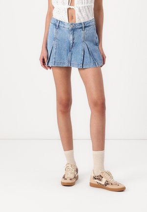 Vrouw draagt een lichtblauwe geplooide denim minirok, witte sokken, beige sneakers met luipaardprint en een wit, knoopcropped topje.