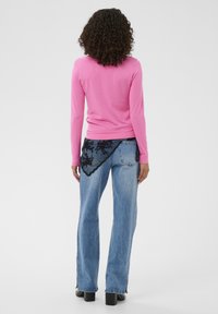 Mujer con pelo rizado, vestida con un top de manga larga rosa brillante y jeans de mezclilla azul claro con detalles de encaje negro, de pie frente a un fondo liso.