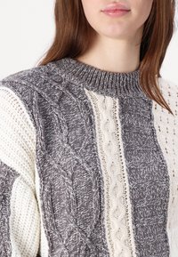 Pull en maille épaisse avec des sections grises et crème, présentant des motifs en câble et des textures. Encolure ras du cou côtelée et détails complexes.