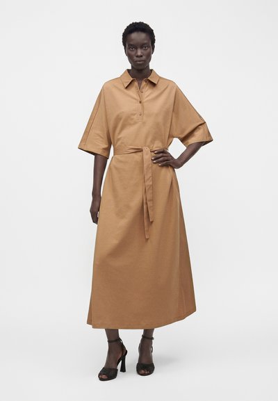 Femme portant une robe midi couleur camel avec un col, une patte de boutonnage, des manches larges, une ceinture à la taille et des sandales noires à talons hauts.