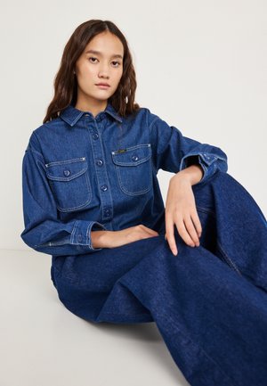 Camicia di denim blu scuro con chiusura frontale a bottoni, due tasche sul petto e dettagli di cuciture visibili; modello seduto con pantaloni abbinati.