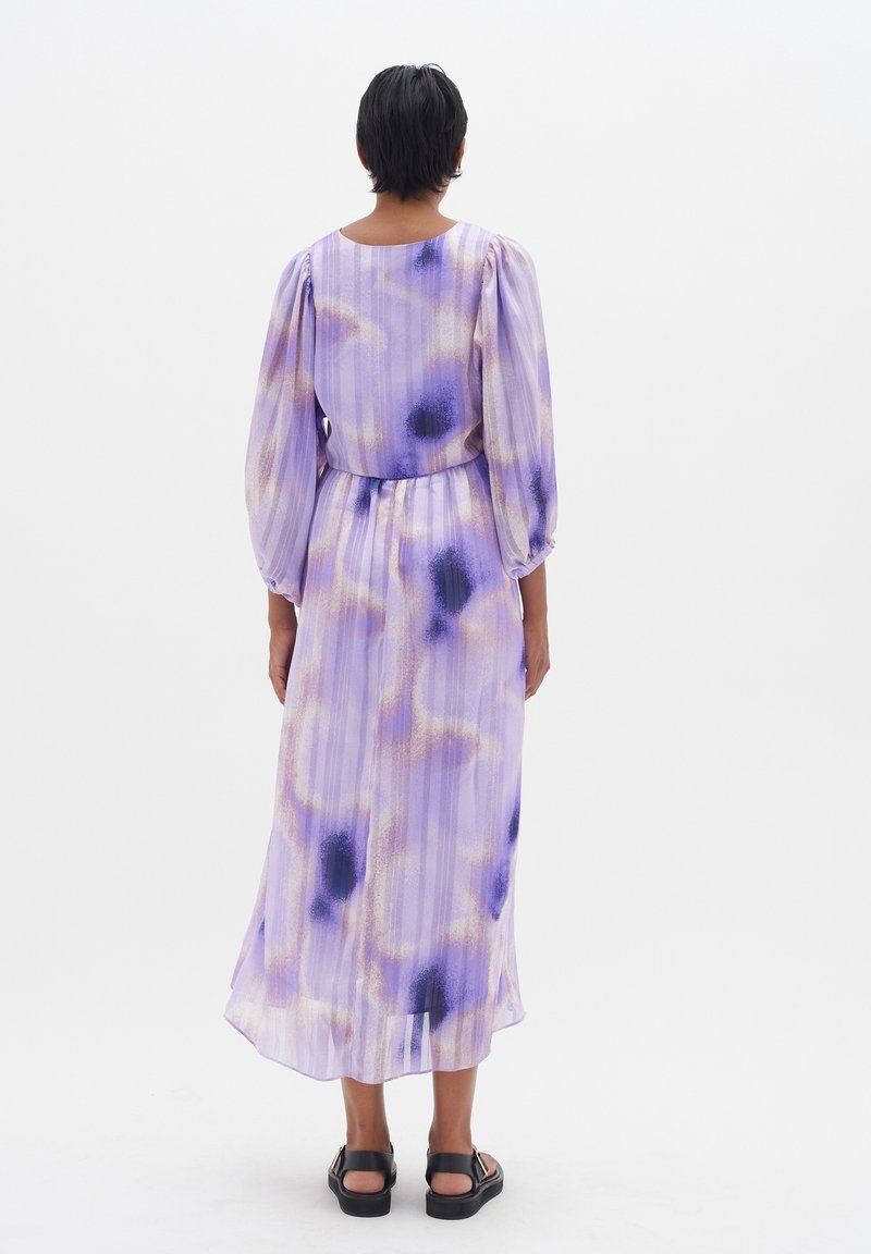 Personne aux cheveux courts noirs portant une longue robe lavande et violette à motifs, avec de longues manches bouffantes et des sandales noires, debout, de dos.