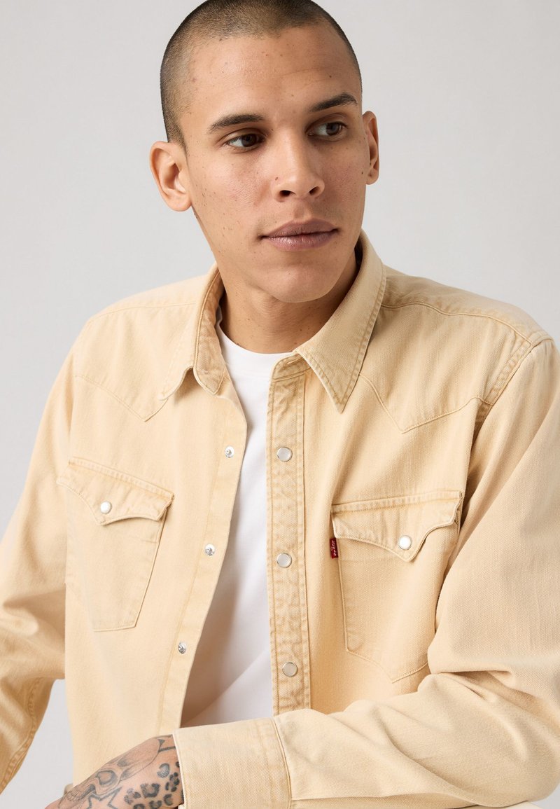 Hombre joven con la cabeza afeitada, vistiendo una camisa de botones beige sobre una camiseta blanca, mirando ligeramente de lado, con un tatuaje visible en el antebrazo.