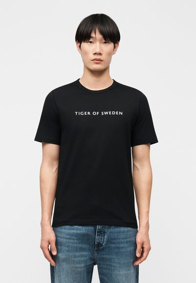 Giovane uomo con capelli neri lisci, che indossa una t-shirt nera "Tiger of Sweden" e jeans blu, in piedi davanti a uno sfondo chiaro e uniforme.