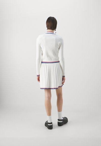 Robe tricotée blanche avec une jupe plissée, bordée de contraste rouge et bleu sur le col et l'ourlet, associée à des chaussures noires.