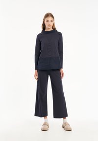 Maglietta a maniche lunghe blu navy con motivo in rilievo "V.5" e un pannello a texture contrastante. Abbinata a pantaloni larghi blu navy e calzature beige.