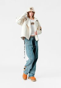 Chaqueta de piel sintética de oveja en crema, camiseta blanca, vaqueros baggy de color azul claro con detalles bordados, botas gruesas naranjas, sombrero de cubo beige con diseño de oso.