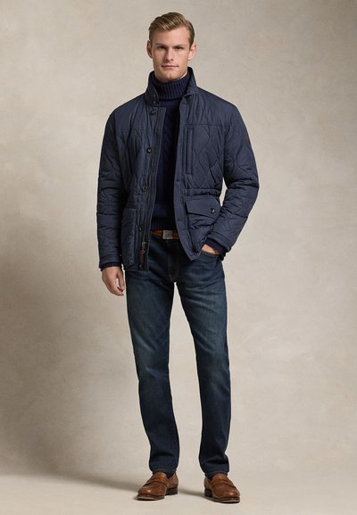 Plavokosi muškarac nosi mornarsko plavu prošivenu jaknu preko mornarsko plavog rolka, tamne traperice i smeđe kožne cipele, stoji s jednom rukom u džepu.