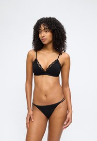Γυναίκα με σγουρά μαλλιά, που φοράει μαύρο σουτιέν με δαντέλα και μαύρο εσώρουχο στυλ μπικίνι, στέκεται μπροστά σε απλό φόντο.