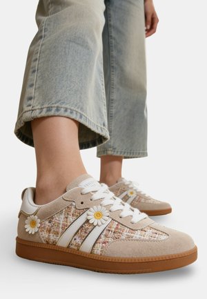Baskets beige et blanches en tissu texturé avec des détails de fleurs de marguerite, portées avec un jean bleu clair raccourci.