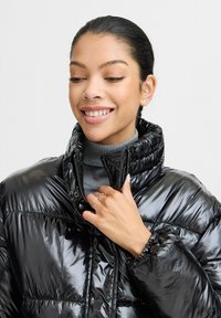 Veste doudoune noire avec finition brillante, col montant, fermeture éclair frontale et poignets élastiques. Portée par-dessus un col roulé gris. Détails de design minimalistes.