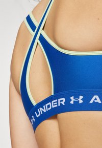 Blue athletic bra with light green accents. Features a racerback design and thick underband with white "UNDER ARMOUR" branding. Smooth texture.