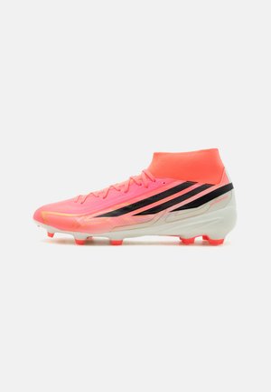Leuchtend korallenfarbener Fußballschuh mit hohem Knöchelkragen, schwarzen diagonalen Streifen, weißer Sohle und orangefarbenen Stollen, seitlich vor weißem Hintergrund dargestellt.