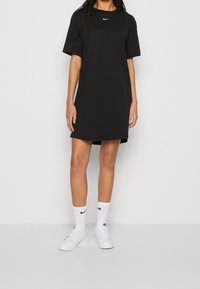 Czarna sukienka T-shirt z krótkim rękawem, uszyta z miękkiego materiału, z białym logo Nike na piersi, w zestawieniu z białymi trampkami i skarpetkami.