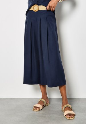 Frau trägt einen marineblauen, plissierten Midirock mit gewebtem, beigem Gürtel und beigen Sandalen mit offenen Zehen und floralen Riemen.