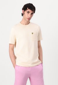 Krämfärgad bomulls-t-shirt med rund hals och korta ärmar, med en liten gul fjäril-logotyp på den vänstra fickan.