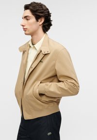 Μπεζ τζάκετ με κολάρο με κουμπιά, φερμουάρ μπροστά και πλαϊνές τσέπες. Διαθέτει κεντημένο πράσινο λογότυπο στο στήθος. Φορά πάνω από ένα ανοιχτόχρωμο πουκάμισο με καρό.