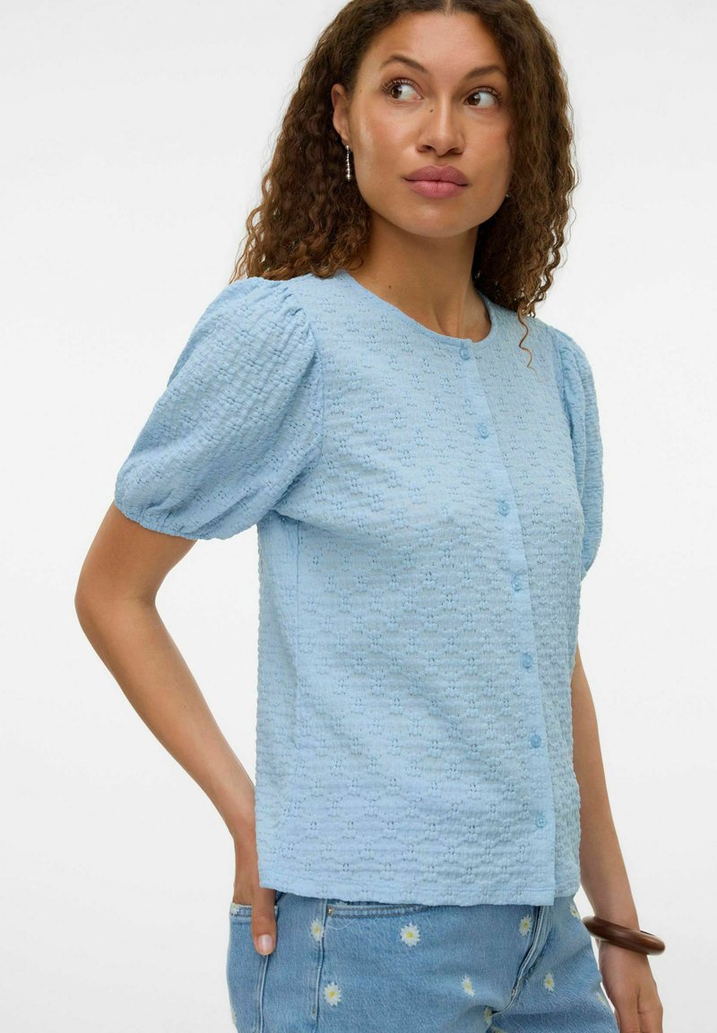 Blusa azzurro chiaro a maniche corte in tessuto testurizzato, con scollatura rotonda e bottoni frontali. Presenta maniche a sbuffo e un orlo dritto.