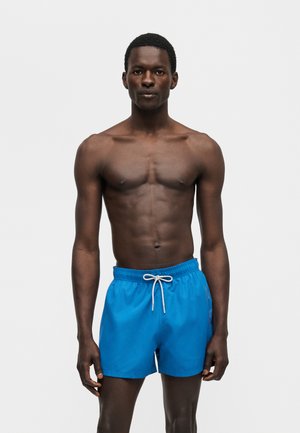 Μαύρος άντρας με κοντά πουα μαλλιά που στέκεται, φοράει φωτεινά μπλε μαγιό με λευκό κορδόνι, σε απλό ανοιχτό φόντο.