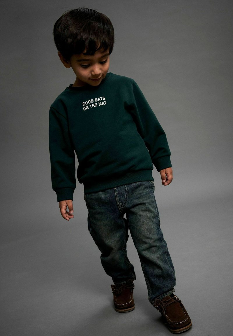 Sweatshirt vert foncé avec le texte "GOOD DAYS ON THE WAY", associé à un jean en denim bleu et des chaussures marron. Design simple, coupe décontractée.