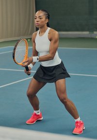 Giocatrice di tennis femminile in una maglietta bianca senza maniche e una gonna nera, con in mano una racchetta arancione, indossando scarpe rosa, su un campo al coperto blu.