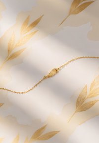 Guldarmband med en vingeformad berlock, med en fin kedja och slät yta, visad mot en ljus bakgrund med blommönster.