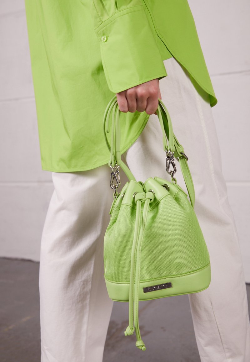 Mano che tiene una borsa a secchiello con coulisse di colore verde lime, indossando una camicia verde lime e pantaloni avorio, su uno sfondo semplice.