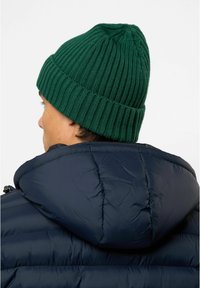 Grønn strikket lue med ribbet tekstur og oppbrett, båret av en person med kort hår, sammen med en mørkeblå dunjakke.