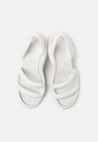 Valkoiset sandaalit paksuilla, kaarevilla remmeillä ja tekstuurilla varustetulla pohjalla. Kevyt rakenne minimalistisella estetiikalla.