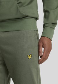 Olivgrüner Hoodie und passende Jogginghosen aus weichem Stoff; mit einem gelben gestickten Logo auf den Hosen. Rippbündchen und -bund.