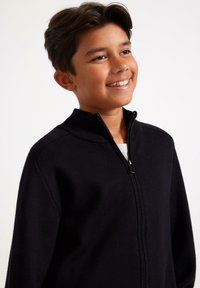 Schwarzer Reißverschluss-Pullover mit hohem Kragen, gestrickter Textur und glatt laufendem Reißverschluss. Über einem grauen Hemd getragen, körperbetontes Design, geeignet für Freizeitmode.