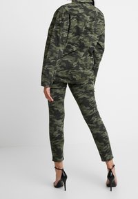 Camo jacket and pants set in green and black, featuring a relaxed fit, buttoned cuffs, and paired with black high-heeled sandals.