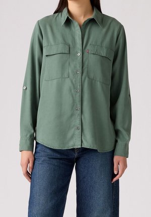 Femme portant une chemise à manches longues boutonnée vert olive avec deux poches poitrine et un jean bleu, debout devant un fond uni.