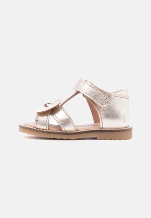 Vaikiška metalinė auksinė sandalė su lipdukinio užsegimo dirželiu prie kulkšnies, atvirais pirštais, dekoratyvine kaspina ir lygiu guminės pado pagrindu ant balto fono.